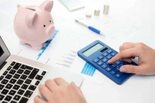 A imagem ilustra elementos fundamentais na gestão de Orçamento Público. Um cofrinho rosa, representando a economia fiscal, é acompanhado por relatórios detalhados, expressos em gráficos e números. Uma das mãos postas no teclado do notebook e a digitando uma calculadora azul-marinho indicam o trabalho preciso de alocação de recursos. Uma caneta azul e torres de moedas dispostas na ordem crescente crescentes completam a composição, ressaltando a importância da transparência e eficiência na administração financeira pública.